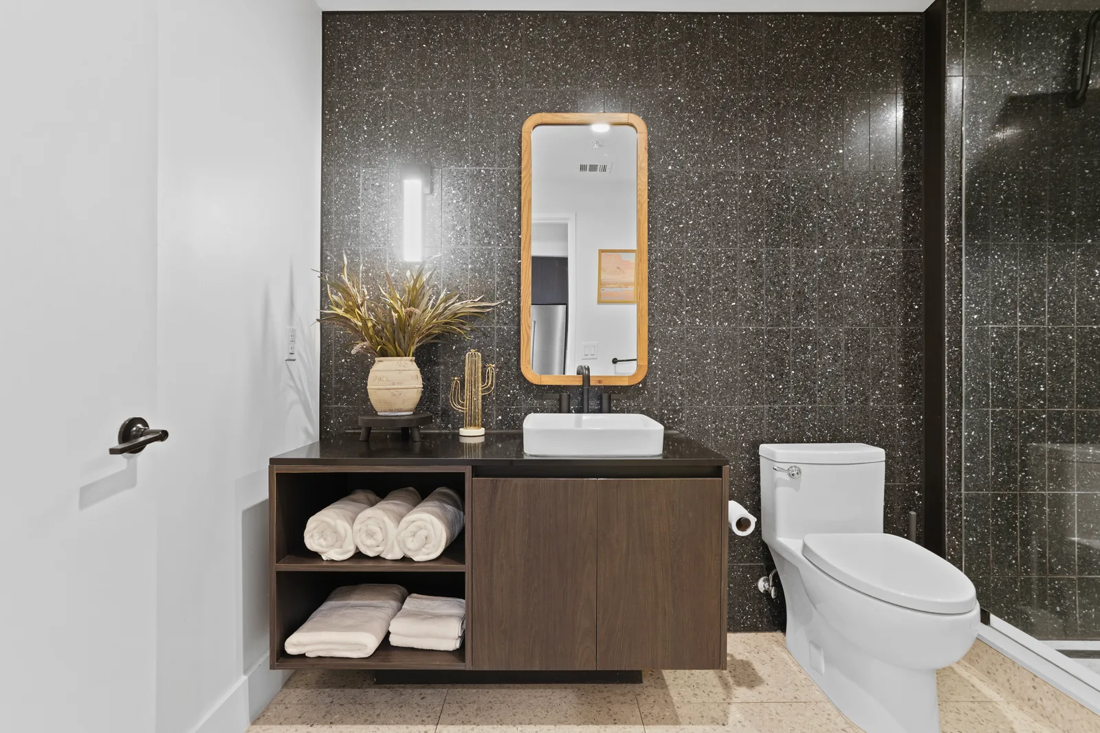 Bathroom with dark speckled tile, wood-framed mirror, floating vanity and rolled white towels