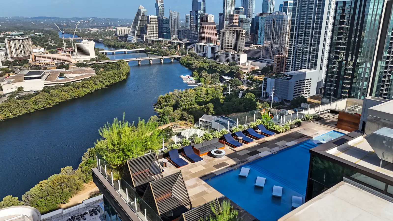 Aerial view of the shared rooftop pool deck with in-pool loungers, lounge chairs and shade cabanas, Lady Bird Lake and the downtown Austin skyline beyond