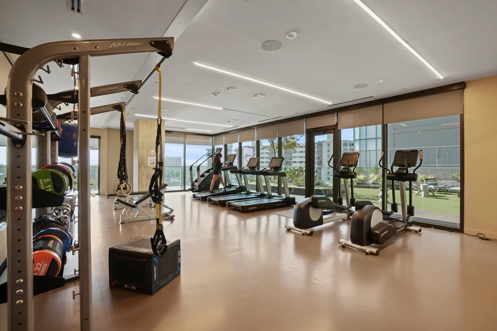 Shared fitness center with a training rig, free weights, treadmills and ellipticals beside floor-to-ceiling windows, with a guest using a treadmill